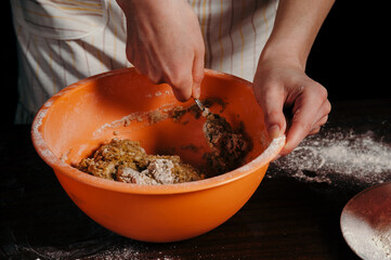 A girl in an apron in a dark kitchen kneads the dough in an orange cup.