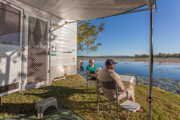 Retired couple sitting outside of caravan with small dog enjoying a wine next to the Lilly filled...
