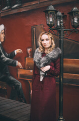 Молодая красивая девушка в бордовом платье и меховом воротнике стоит под фонарем на фоне граффити пианиста