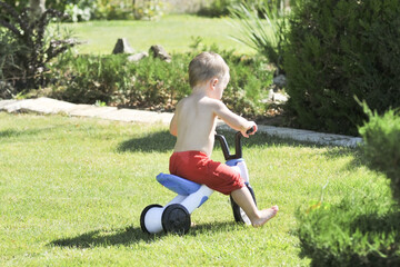Fototapeta premium Little boy pours from a hose in the garden on a hot summer day on a green lawn, splashing water