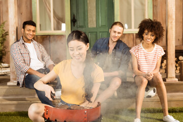 young woman preparing barbecue grill on patio while her friends sitting at porch