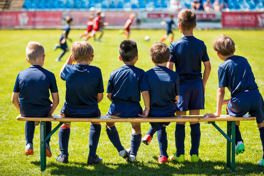 Soccer Match For Children. Young Boys Playing Tournament Soccer Match. Youth Soccer Club Footballers. Young Football Players in Blue Jerseys. Young Soccer Team Sitting on Wooden Bench.
