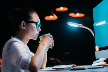Side view of young businesswoman in eyeglasses holding cup while working late in office