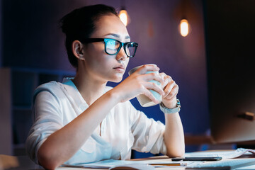 Young businesswoman in eyeglasses holding cup while working late in office