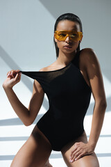 african american stylish model in black swimsuit and protective goggles posing on grey and looking...
