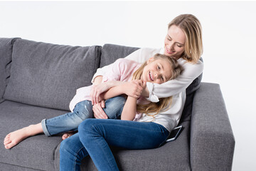 Fototapeta na wymiar smiling mother with little daughter sitting on sofa together isolated on white
