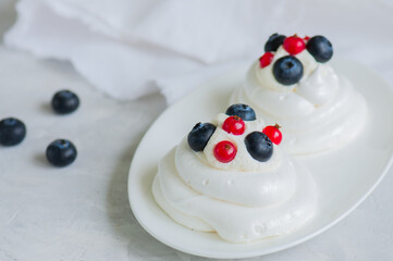 Close up of homemade individual meringue cakes Pavlova with whipped cream with mascarpone fresh blueberries and red currants in a white plate on a white stone background. Copy space.