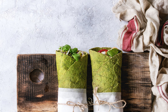Green spinach matcha tortillas wrapped in paper with ingredients above. Sweet corn, avocado, basil, sprouts, mushrooms served on wood board over gray texture background and textile. Flat lay