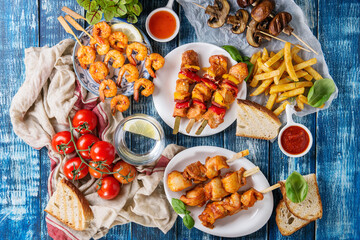 Fototapeta na wymiar Variety of BBQ snack lunch. Plates grilled spicy prawn kebabs, chicken, pork, vegetables, mushrooms skewers, bread, french fries potatoes with sauces and greens over blue wooden background. Flat lay