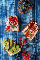 Variety of dessert sandwiches with berries and cream cheese and chocolate swirl. Red currant, blueberries, sliced kiwi, figs over blue wooden plank background. Flat lay, summer appetizer concept