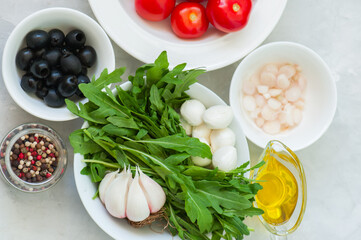 Ingredients for healthy salad. Tomatoes, rucola, onion, mozzarella, olives, garlic and pepper with olive oil on a white stone background.