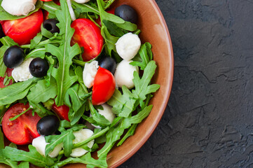 Close up of fresh salad with rucola, tomatoes, olives, onion, mozzarella and spices in a bowl on a black slate background with copy space. Healthy food, Diet, Detox, Clean Eating or Vegetarian concept