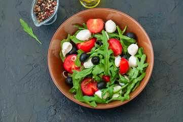 Close up of fresh salad with rucola, tomatoes, olives, onion, mozzarella and spices in a bowl on a black slate background with copy space. Healthy food, Diet, Detox, Clean Eating or Vegetarian concept