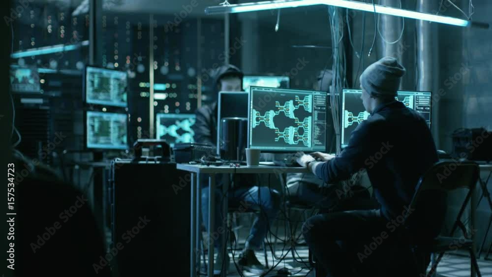 Poster hooded hacker takes his place in at his desk in hacker's den. place is neon, dark and has many displ
