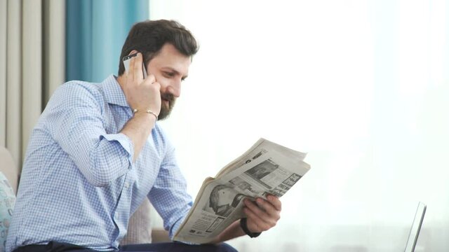 A stylish man with a beard in bright and cozy living room, holds a newspaper in his hands and speaks on the phone. An attractive brutal man with a beard and in a blue shirt makes a working phone call.