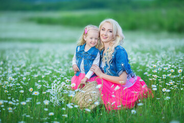 Fototapeta premium Mom and daughter on a picnic in the chamomile field. Two beautiful blondes wearing jeans jacket and pink skirt Mother and baby girl embracing in the chamomile field with basket of bouquet dandalion