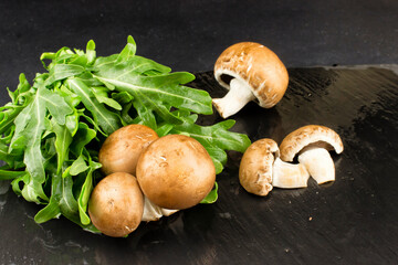 Fresh raw mushrooms brown champignons and green arugula on a dark background in a low key