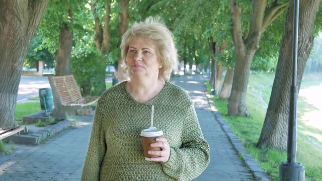 Elderly woman walking park lane drinking coffee. Senior female walking in the park