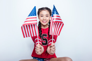 smiling asian girl in red sweatshirt sitting and holding little american flags isolated on grey, 4th july - Independence Day
