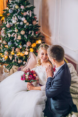 Bride in beautiful dress and groom in gray suit sitting on sofa indoors in white studio interior like at home. Newlyweds kiss each other at home. Flowers bouquet in woman hand.