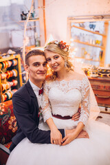 Bride in beautiful dress and groom in black suit sitting on sofa indoors in white studio interior like at home. Trendy wedding style. Groom.bride hugging