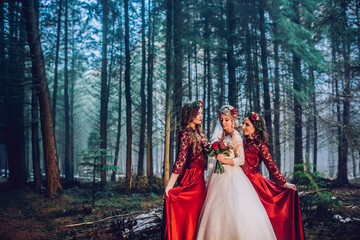 Fototapeta premium Bride and bridesmaids in red dresses stand on the path in pine forest.