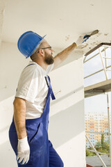 Construction worker in work attire, protective gloves and a helmet on site. Remove the old paint spatula from the ceiling.