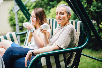 Smiling middle-aged mother and adult daughter in law sitting on the sofa and drinking tea at yard.