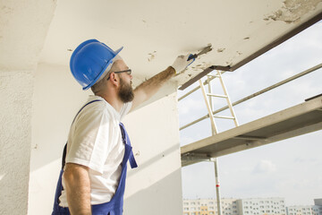 Construction worker in work attire, protective gloves and a helmet on site. Remove the old paint spatula from the ceiling. © Tomasz