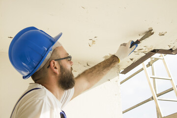 Construction worker in work attire, protective gloves and a helmet on site. Remove the old paint spatula from the ceiling. © Tomasz