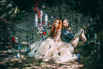 Beautiful young couple together near dark circle arch with flowers. Man with tattoo and long hair with beard. Woman with red hair. Fairytale concept. - obrazy, fototapety, plakaty