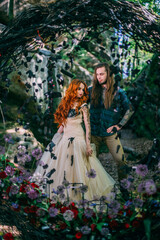 Obraz na płótnie Canvas Beautiful young couple together near dark circle arch with flowers. Man with tattoo and long hair with beard. Woman with red hair. Fairytale concept.