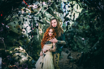 Fototapeta na wymiar Beautiful young couple together near dark circle arch with flowers. Man with tattoo and long hair with beard. Woman with red hair. Fairytale concept.