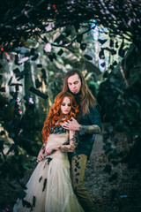 Fototapeta na wymiar Beautiful young couple together near dark circle arch with flowers. Man with tattoo and long hair with beard. Woman with red hair. Fairytale concept.