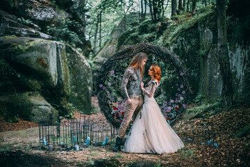 Beautiful young couple together near dark circle arch with flowers. Man with tattoo and long hair with beard. Woman with red hair. Fairytale concept.