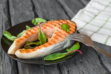 View at the grilled steak of salmon fish with fork on a black plate and green leaves of chard salad. Dish on the black wooden table. White checkered napkin at the background.