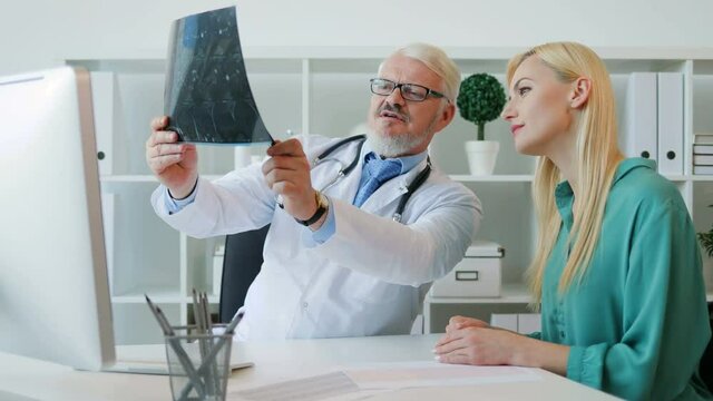 Midle aded caucasian senior doctor man sitting in his offise, showing to female patient x-ray image and talking to patient about it. Indoor.