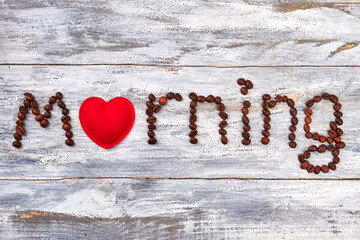 Textile heart between coffee beans. Text morning on wooden table.