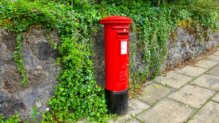 Fototapeta na wymiar Lennoxtown,Scotland, 20 May 2017: Red Post Office pillar box in front of stone wall covered in ivy. 