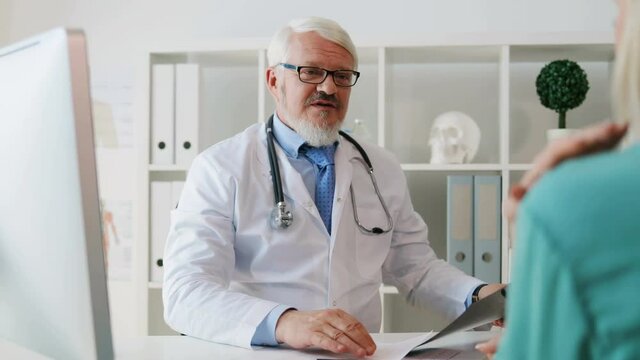 Caucasian middle aged doctor in glasses with beard looking roentgen image, explaining something patient. Indoor.