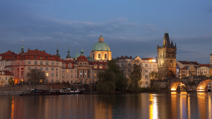 Fototapeta na wymiar Old Town Bridge Tower and Vltava river bank with Charles bridge after dark. The dome of St. Francis Of Assisi Church in the background. Docked boats of Old Town river bank of Vltava.