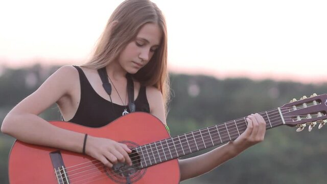 Young woman outdoors softly strumming her guitar. Teen girl learning to play acoustic guitar in nature