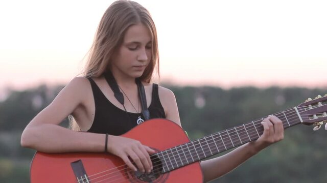 Teenage girl practicing guitar then smiling to camera at sunset outdoors