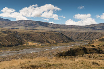 Fototapeta na wymiar Middle Earth, New Zealand - March 14, 2017: High desert landscape with Lake Clearwater overflow drain to Rangitata River under blue sky with white clouds. Set in brown dry vegetation on mountains.