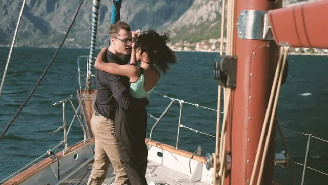 Loving happy couple dancing on yacht walk on lake outdoors. Beautiful woman with dark skinned hair black skirt blue topic smiling hugs man in shirt trousers looks at wife leads in dance. expensive
