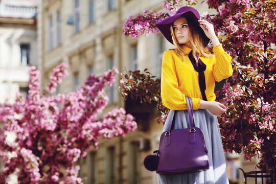 Outdoor portrait of young beautiful girl posing in street. Model wearing stylish hat, shirt, skirt, holding purple bag, handbag. City lifestyle. Female fashion concept. Copy, empty space for text
