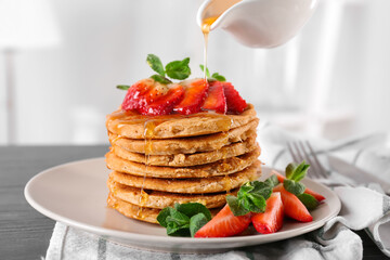 Pouring sweet sauce from gravy boat onto stack of delicious coconut pancakes with strawberry and mint