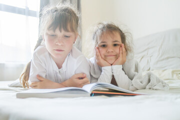 indoor portrait of young european girls - two sisters - lying in bed and reading a book