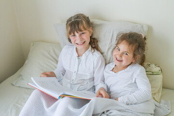indoor portrait of young european girls - two sisters - lying in bed and reading a book