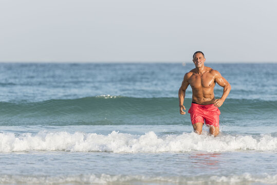 Mature older muscular fit Arab Male running out of the ocean on the beach, shirtless, wearing red shorts 